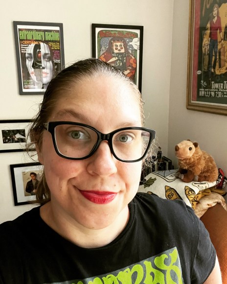 Anna, a white nonbinary person wearing glasses and a black t-shirt, stands in front of several concert posters.