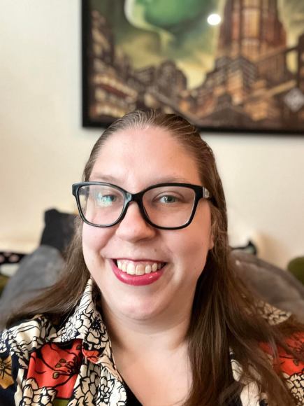 Photo of Anna, a white nonbinary person, smiling. They wear glasses and a floral print button down shirt.
